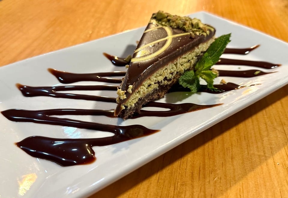 Slice of chocolate pistachio cake with chocolate drizzle and mint garnish on a white plate.