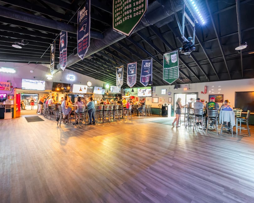 A lively bar interior with patrons seated at a long bar, sports banners hanging from the ceiling.