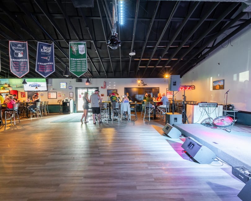 A lively bar interior with patrons at high-top tables and a stage with musical equipment.