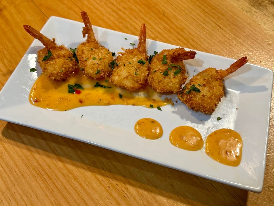 Five golden-brown coconut shrimp with tails on, drizzled with orange sauce and parsley on a white plate.