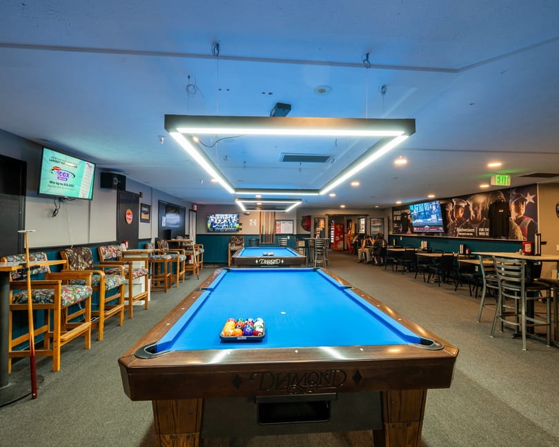 A spacious game room with two blue-felt pool tables, high-top seating, and TVs on the walls.