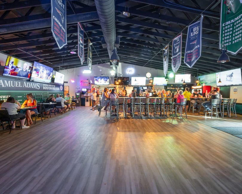 A lively sports bar interior with patrons at tables and a long bar, surrounded by TVs and sports banners.